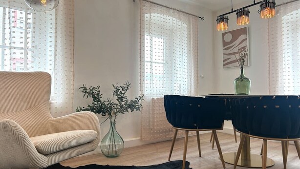 Ein Wohnzimmer mit einer Couch, einem Esstisch mit Stühlen, einer Vase mit Pflanzen und einem schwarzen Teppich.