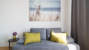 Un divano grigio con due cuscini gialli, un tavolino rotondo con un vaso di fiori viola e un quadro a tema spiaggia sulla parete.