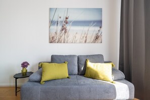 Ein graues Sofa mit zwei gelben Kissen, ein kleiner runder Tisch mit einer Vase voller lila Blumen und ein Strandbild an der Wand.