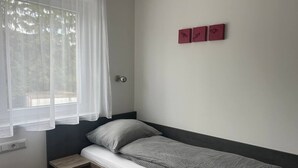 A single bed with a grey patterned bedspread and pillows, a small wooden nightstand, a window with sheer curtains, and three red wall decorations. - FarawayHomes Aparthotel Kremsmünster #13 (Kremsmünster)