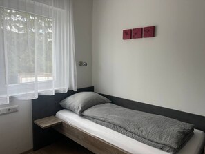 Ein Einzelbett mit grau gemustertem Bettbezug, ein Nachttisch aus Holz, ein Fenster mit durchscheinenden Vorhängen und drei rote Wanddekorationen.