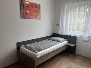 Ein Einzelbett mit Holzrahmen, ein Nachttisch, ein Fenster mit durchsichtigen Vorhängen und ein an der Wand hängendes abstraktes Gemälde.