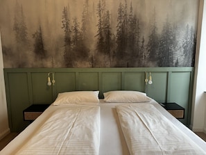 En dubbelsäng med två sängbord, en grön sänggavel och en väggmålning med skogsmotiv.