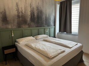 Ein Doppelbett mit weißer Bettwäsche, ein Kopfteil, ein Nachttisch und eine Wand mit einem Waldgemälde.