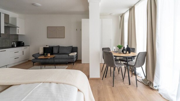 Quarto de hotel moderno com cama, sofá, mesa de jantar com cadeiras, cozinha e uma grande janela com cortinas.
