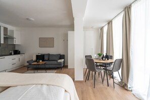 Moderni hotellihuone, jossa on sänky, sohva, ruokapöytä tuoleineen, keittiö sekä suuri ikkuna, jossa on verhot.