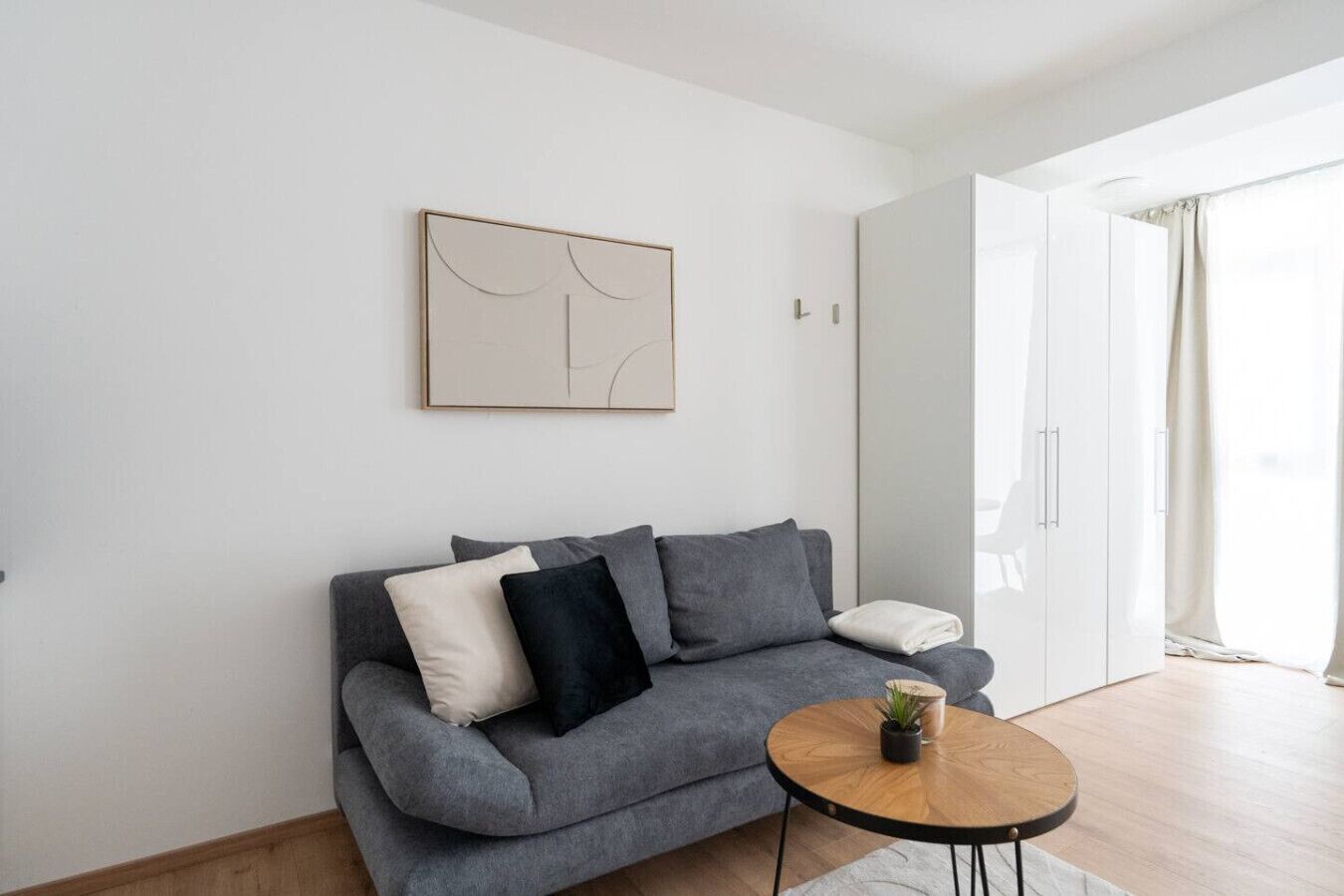 Un saló modern amb un sofà gris, una taula rodona petita amb una planta i una obra d'art minimalista a la paret.