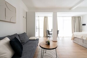 Quarto moderno e bem iluminado com sofá cinzento, mesa de centro redonda com uma planta e uma cama ao fundo.