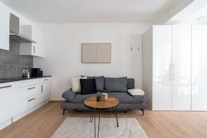 Ein modernes Wohnzimmer mit grauem Sofa, einem runden Holztisch und einer Küchenzeile mit weißen Schränken und einer grauen Arbeitsplatte.