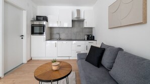 A compact kitchen with white cabinets, a stove, and a sink. There's a gray sofa with a plant on a wooden table.