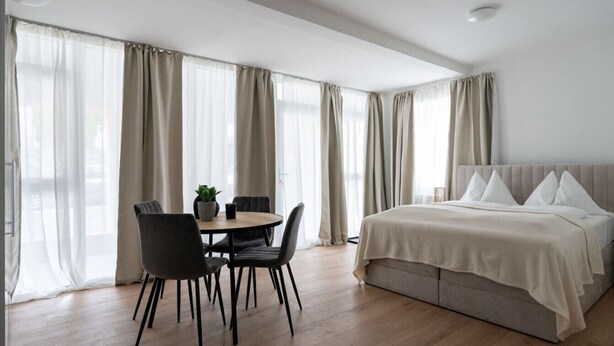 Ett modernt hotellrum med en säng, ett matbord med stolar och stora fönster med gardiner.
