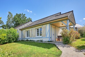 A two-story house with a covered porch, a small garden, and a clear blue sky. - Stunning Bungalow With Pool for 6 (Branville)