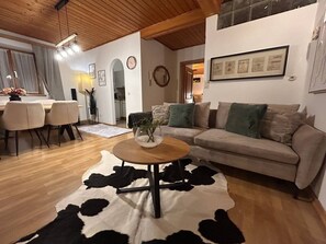 Ein Wohnzimmer mit rundem Holztisch, einer Couch mit Kissen, einem Essbereich mit Stühlen und einer durch einen Bogen sichtbaren Küche.