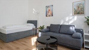 Una habitación moderna con un sofá gris, una mesita redonda pequeña, una cama con ropa de cama blanca y dos cuadros enmarcados en la pared.