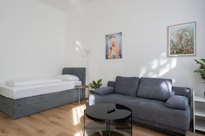 Une chambre moderne avec un canapé gris, une petite table ronde, un lit avec une literie blanche et deux tableaux encadrés au mur.