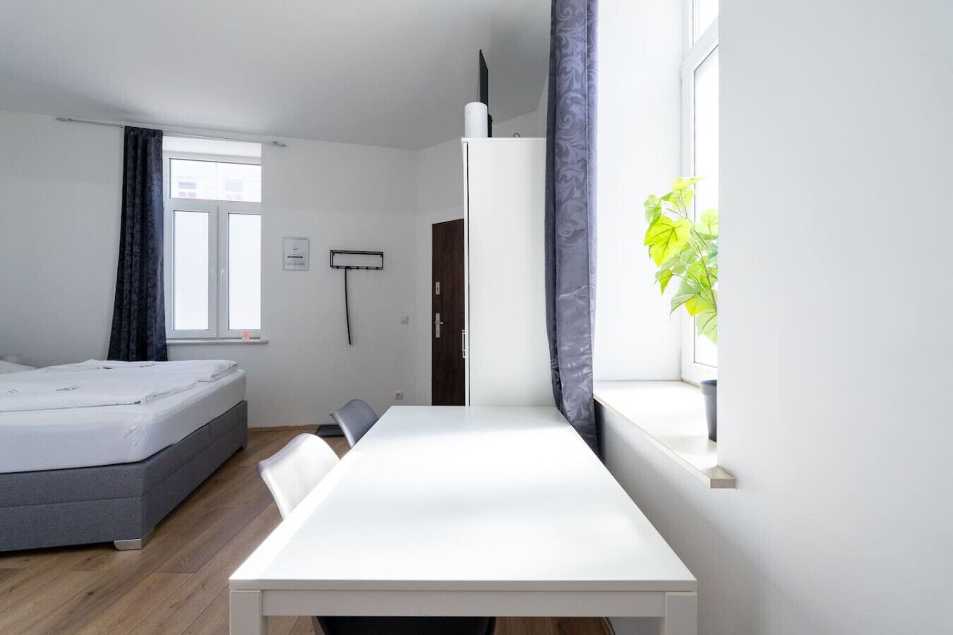 A modern, well-lit room with a bed, a dining table, and a window with curtains.