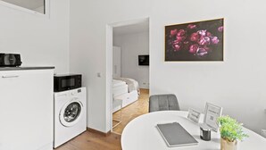Ett kompakt bostadsutrymme med ett litet kök, en tvättmaskin, en säng, en matplats och ett inramat konstverk med blommotiv.