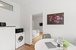Un espacio habitable pequeño con kitchenette, lavadora, cama, zona de comedor y una obra de arte floral enmarcada.