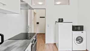 Ett kompakt kök med spishäll, mikrovågsugn och tvättmaskin, en dörr som leder till ett annat rum och infällda taklampor.