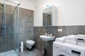 Ein modernes Badezimmer mit Duschkabine aus Glas, Toilette, Waschbecken mit Spiegel sowie gestapelter Waschmaschine und Trockner.