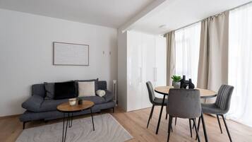 Een moderne woonkamer met een grijze bank, een ronde tafel en stoelen, een klein vloerkleed en een lijst met een foto aan de muur.