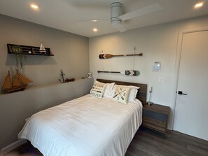 A bedroom with a bed, a ceiling fan, a nightstand, and wall-mounted decorations including a sailboat model and wooden paddles.