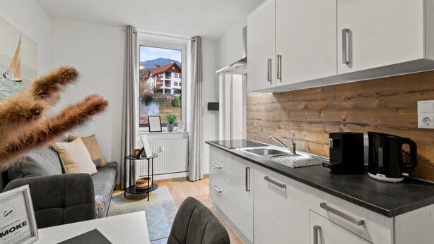 A modern kitchen with white cabinets, a wooden accent wall, a coffee maker, and a view of a residential area. - FarawayHomes Studio Mürzzuschlag #4 (Mürzzuschlag)