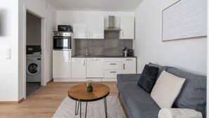 Un espacio habitable compacto con kitchenette, un sofá gris, una mesita redonda con una planta y una lavadora al fondo.