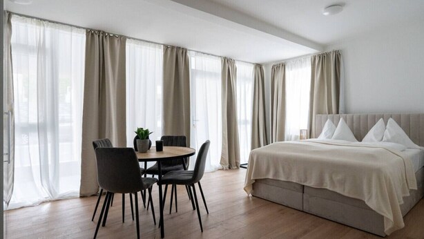 Una habitación de hotel moderna con una cama, una mesa de comedor con sillas y ventanas grandes con cortinas.