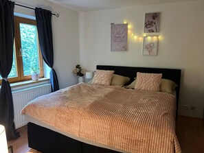 Ein Schlafzimmer mit Bett, Fenster mit Vorhängen, Wandbild und Nachttisch mit Lampe.