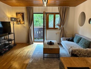 Ein Wohnzimmer mit Sofa, Couchtisch, TV-Schrank und Blick auf Grünflächen.