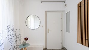 Un corridoio con una porta bianca, uno specchio rotondo, un tavolino con un vaso e un pannello di legno al muro.
