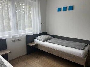 Ein kleines, modernes Zimmer mit einem Einzelbett, einem Fenster mit leichtem Vorhang und einem an der Wand befestigten Regal.