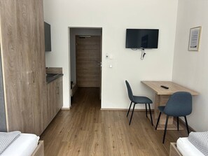 Ein modernes Zimmer mit Holzboden, einem Schreibtisch mit zwei Stühlen, einem an der Wand befestigten Fernseher und einer Tür zu einem weiteren Raum.