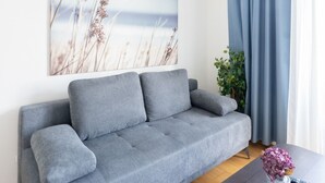A grey sofa, a painting of tall grasses, a blue curtain, a small plant, and a remote control on a dark table.