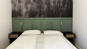Una cama doble con cabecera verde, dos mesitas de noche con lámparas y un mural temático de bosque.