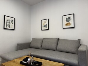 Un sofá gris con tres cojines, una mesa de centro de madera con una botella y un vaso, y tres obras de arte enmarcadas en la pared.