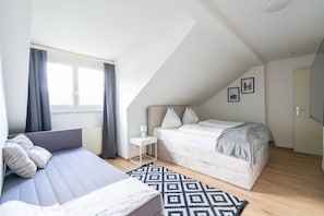 Un dormitorio con cama, sofá, una mesita de noche, ventana con cortinas, alfombra estampada y una puerta.