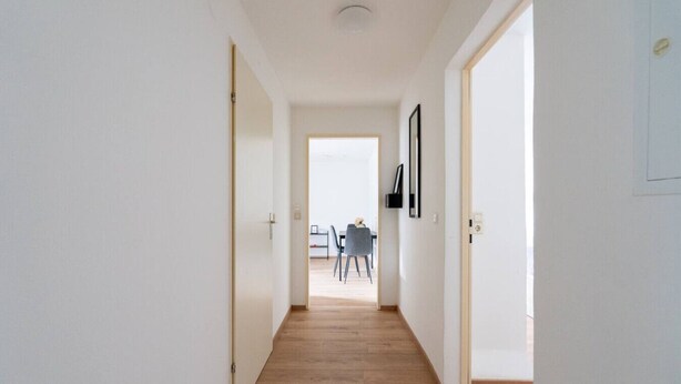 Un pasillo con suelo de madera, paredes blancas y una puerta que da a una habitación con una mesa y sillas.