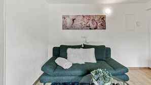 A green sofa with a white pillow and a rolled white towel, a glass table with a plant, and a floral painting on the wall. - FarawayHomes Studio Leoben #P81 (Donawitz)