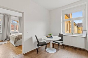 Una habitación con cama, una mesa redonda con dos sillas, un ventanal con vistas a edificios y un radiador.