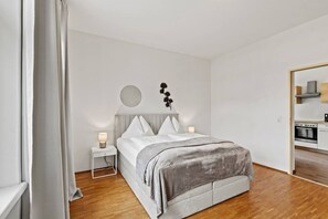 A modern bedroom with a bed, bedside tables, a wall-mounted light, and a kitchen area visible through an open door. - FarawayHomes Studio Leoben #K7 (Donawitz)