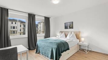 Een slaapkamer met een bed, nachtkastje en uitzicht op gebouwen door een raam.