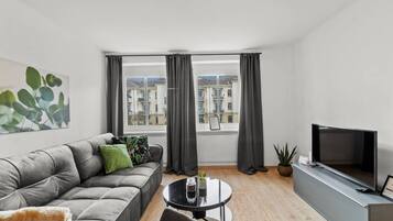 Een moderne woonkamer met een grijze bank, een kleine ronde tafel, een televisie en een groot raam met gordijnen.