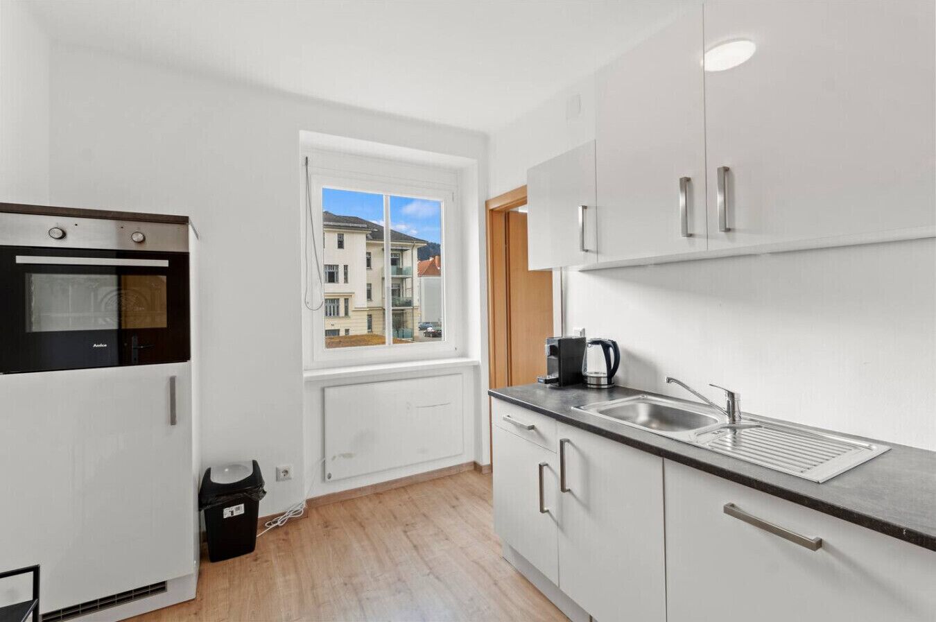 Een moderne keuken met een ingebouwde oven, een raam met uitzicht op gebouwen, een spoelbak en kastjes.