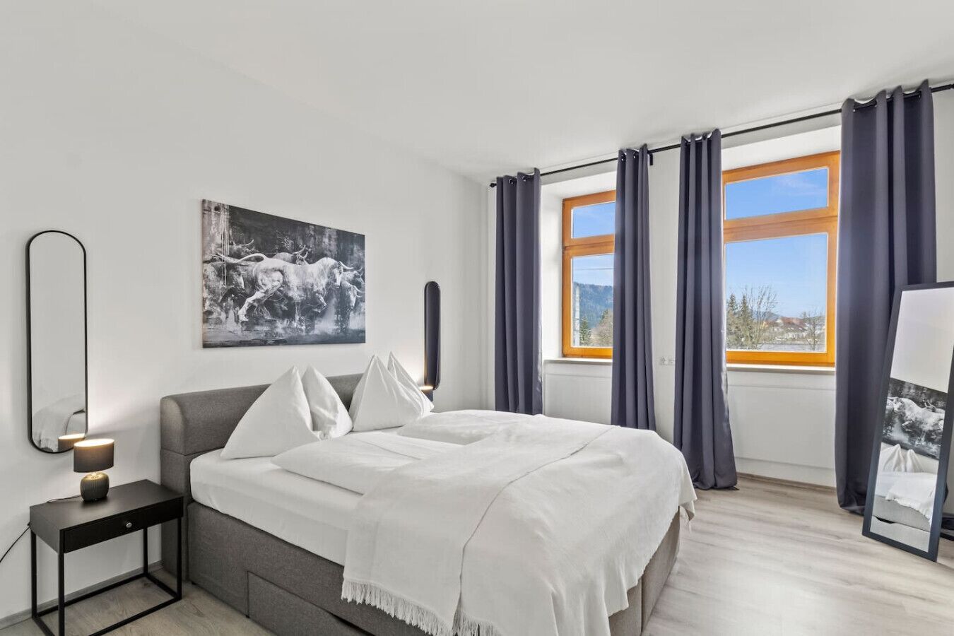 A modern bedroom with a bed, a bedside table with a lamp, a large window with curtains, and a wall-mounted mirror.. FarawayHomes Studio St. Michael #5