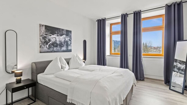 Un dormitorio moderno con una cama, una mesita de noche con lámpara, un ventanal con cortinas y un espejo montado en la pared.