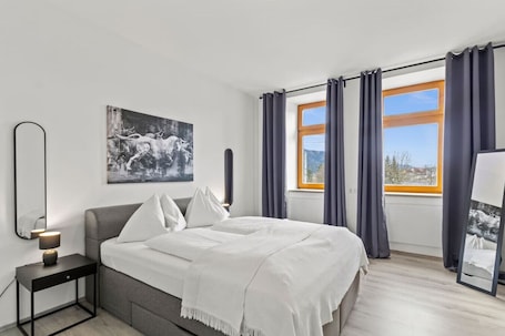 A modern bedroom with a bed, a bedside table with a lamp, a large window with curtains, and a wall-mounted mirror.. FarawayHomes Studio St. Michael #5