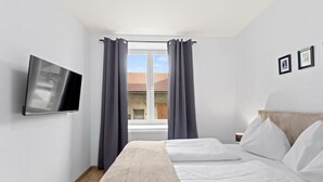 Ein Schlafzimmer mit einem Bett, einem an der Wand befestigten Fernseher und einem Fenster mit Vorhängen.