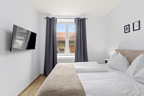 Ein Schlafzimmer mit einem Bett, einem an der Wand befestigten Fernseher und einem Fenster mit Vorhängen.